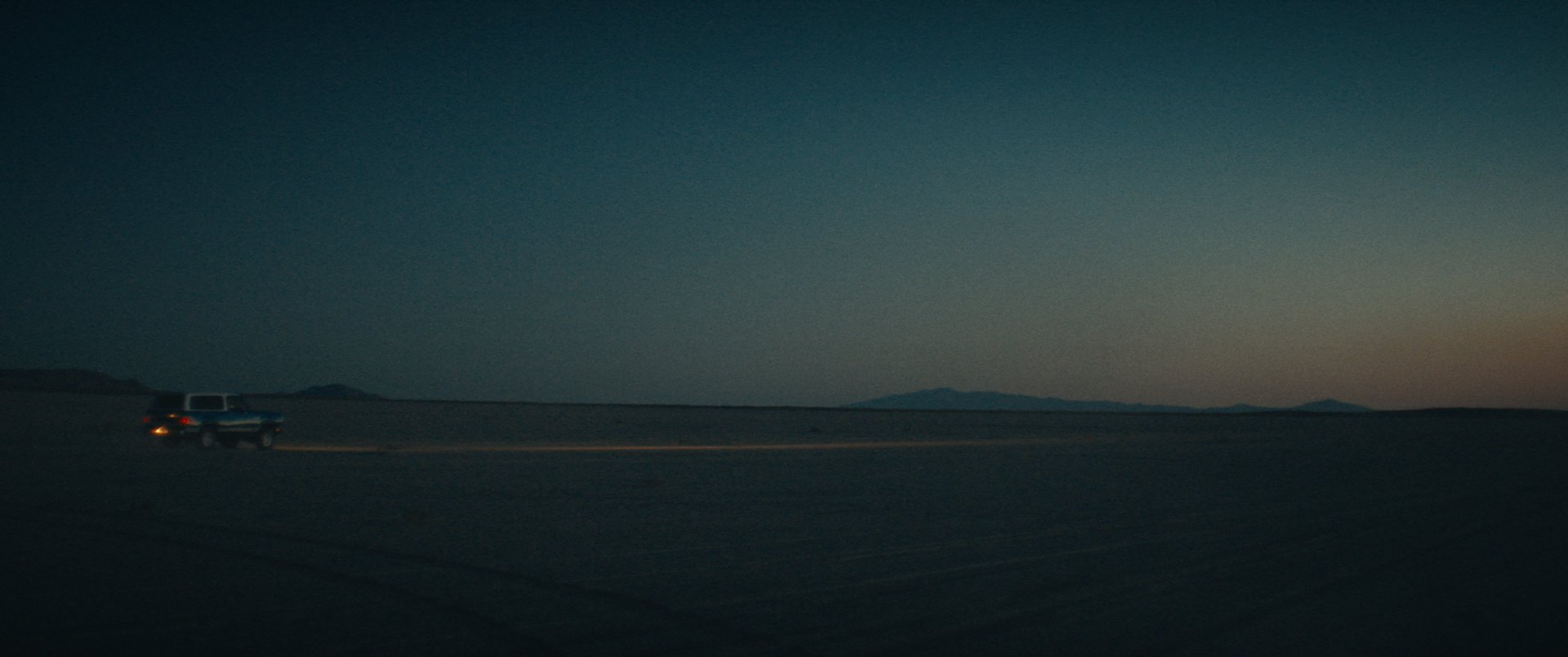 A lone truck crosses a desert salt flat at dusk under a deep teal sky